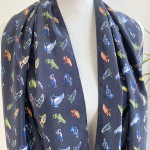 May include: A navy blue scarf with a repeating pattern of colorful frog illustrations. The scarf is draped over a white mannequin, showcasing its design and texture. The frogs are in various colors, including green, blue, red, and yellow.