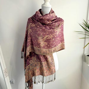 May include: A burgundy paisley patterned scarf with a decorative border and black fringe. The scarf is draped over a white mannequin. The scarf is made of a soft material and is suitable for cool weather.