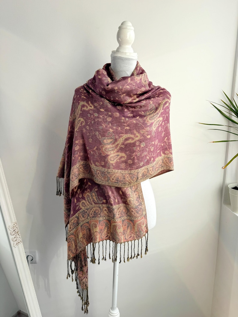 May include: A burgundy paisley patterned scarf with a decorative border and black fringe. The scarf is draped over a white mannequin. The scarf is made of a soft material and is suitable for cool weather.