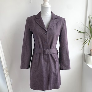 May include: A plum-coloured coat with a belted waist and three buttons. The coat is displayed on a white mannequin. The coat has a notched collar and long sleeves. The coat is made of a textured fabric.
