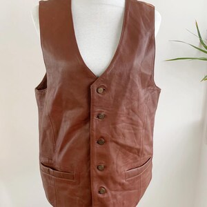 May include: A brown leather vest with a V-neck and a button-up front. The vest has two front pockets and is displayed on a mannequin. The vest is a medium brown color.