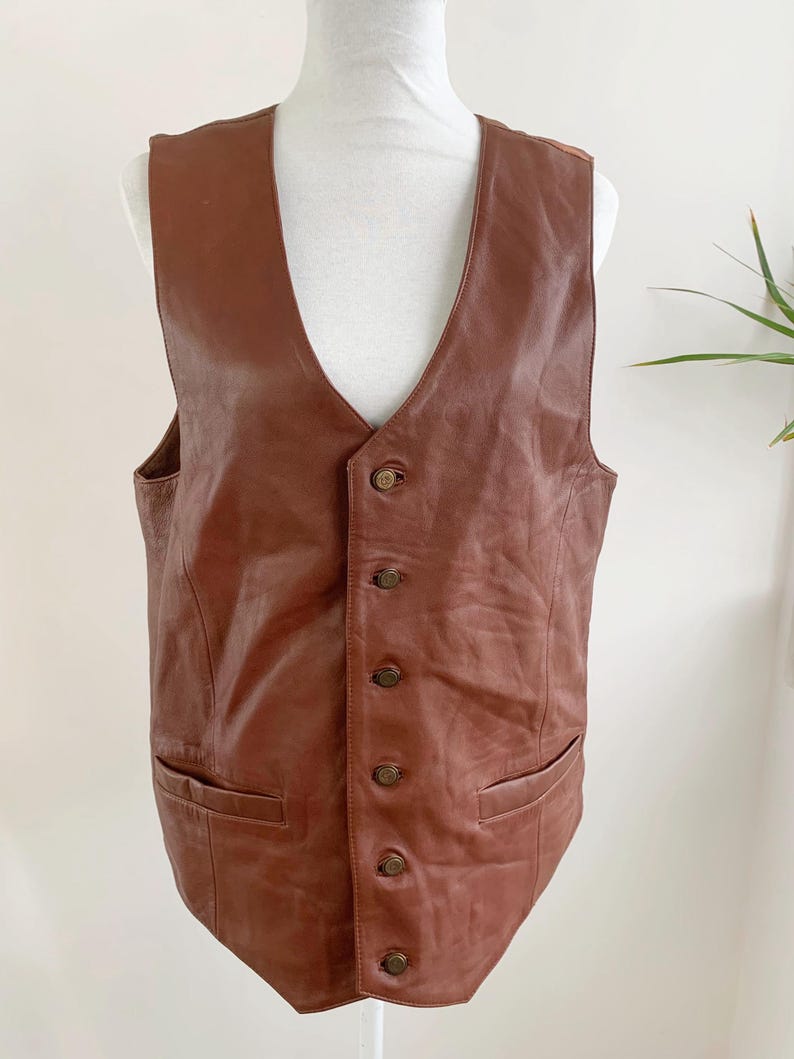 May include: A brown leather vest with a V-neck and a button-up front. The vest has two front pockets and is displayed on a mannequin. The vest is a medium brown color.