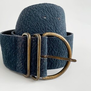 May include: A wide, dark blue belt with a textured finish. It has a large, antique brass buckle with a single prong. The belt is rolled up, displaying its width and the buckle's design.