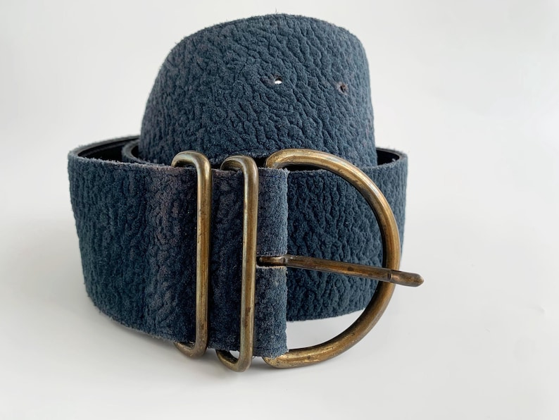 May include: A wide, dark blue belt with a textured finish. It has a large, antique brass buckle with a single prong. The belt is rolled up, displaying its width and the buckle's design.