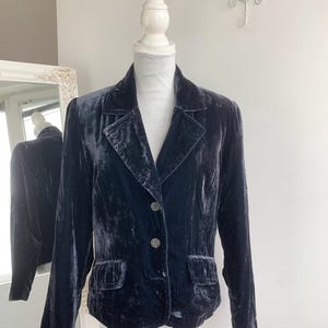 May include: A dark blue velvet blazer displayed on a mannequin. The blazer has a classic design with a notched collar, two front pockets, and two buttons. The fabric has a soft, lustrous appearance.
