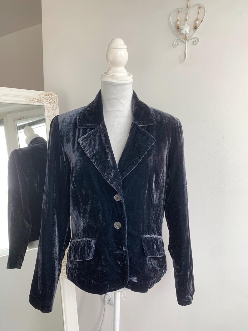 May include: A dark blue velvet blazer displayed on a mannequin. The blazer has a classic design with a notched collar, two front pockets, and two buttons. The fabric has a soft, lustrous appearance.