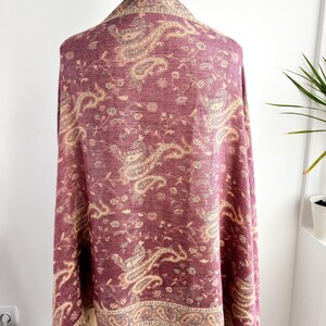 May include: A burgundy paisley shawl draped over a mannequin. The shawl features a detailed pattern in beige and gray, with a decorative border and fringe. The fabric is soft and flowing, perfect for adding a touch of elegance.