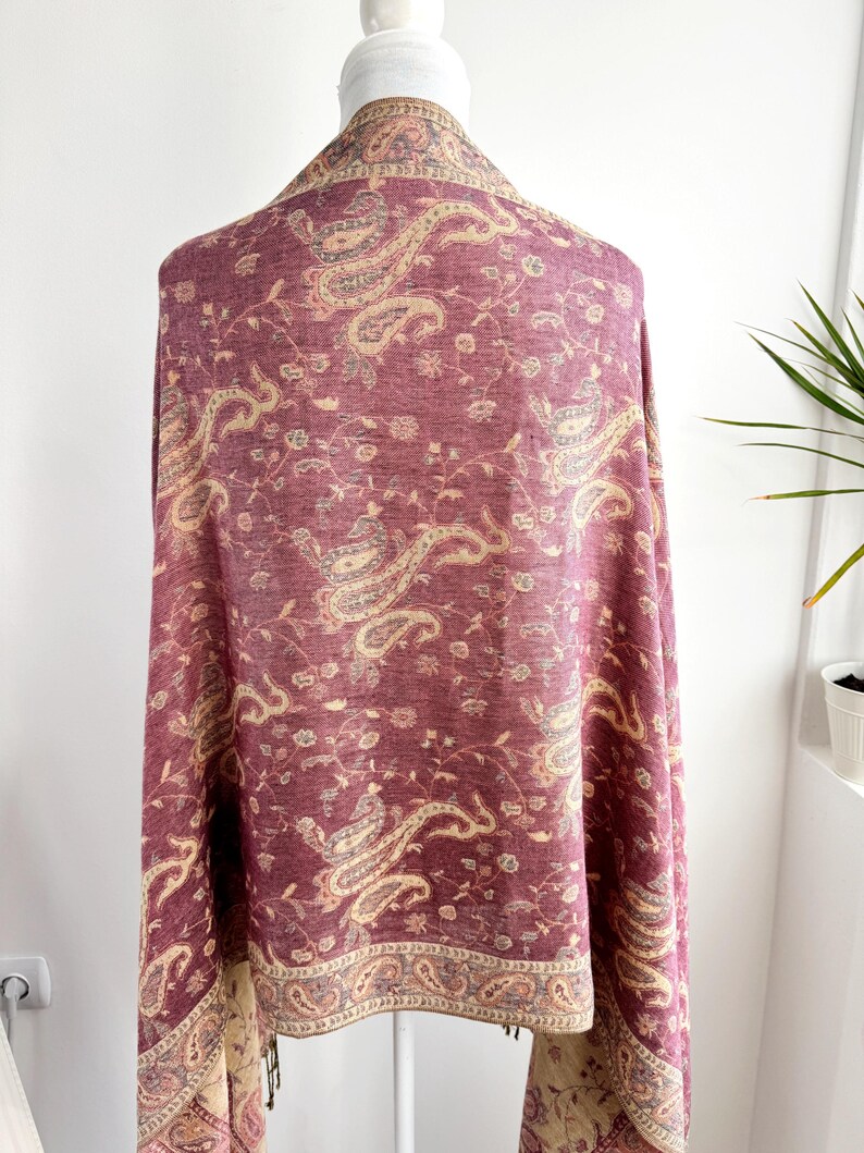 May include: A burgundy paisley shawl draped over a mannequin. The shawl features a detailed pattern in beige and gray, with a decorative border and fringe. The fabric is soft and flowing, perfect for adding a touch of elegance.
