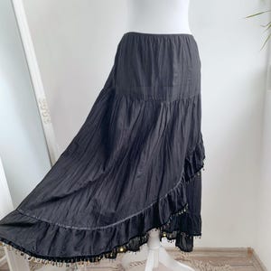 Può includere: Gonna lunga nera con un design a balze a strati. La gonna ha una silhouette fluida ed è adornata da piccoli abbellimenti color oro lungo l'orlo. La gonna è esposta su un manichino bianco.