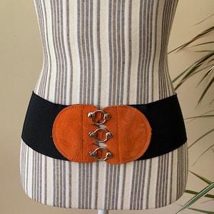 Könnte beinhalten: Ein schwarzer Gummibandgürtel mit einer braunen, ovalen Schnalle aus Wildleder mit vier silbernen Metallringen.