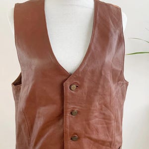 May include: A brown leather vest with a V-neck and four decorative buttons. The vest has two front pockets and is displayed on a white mannequin. The leather has a slightly distressed appearance.