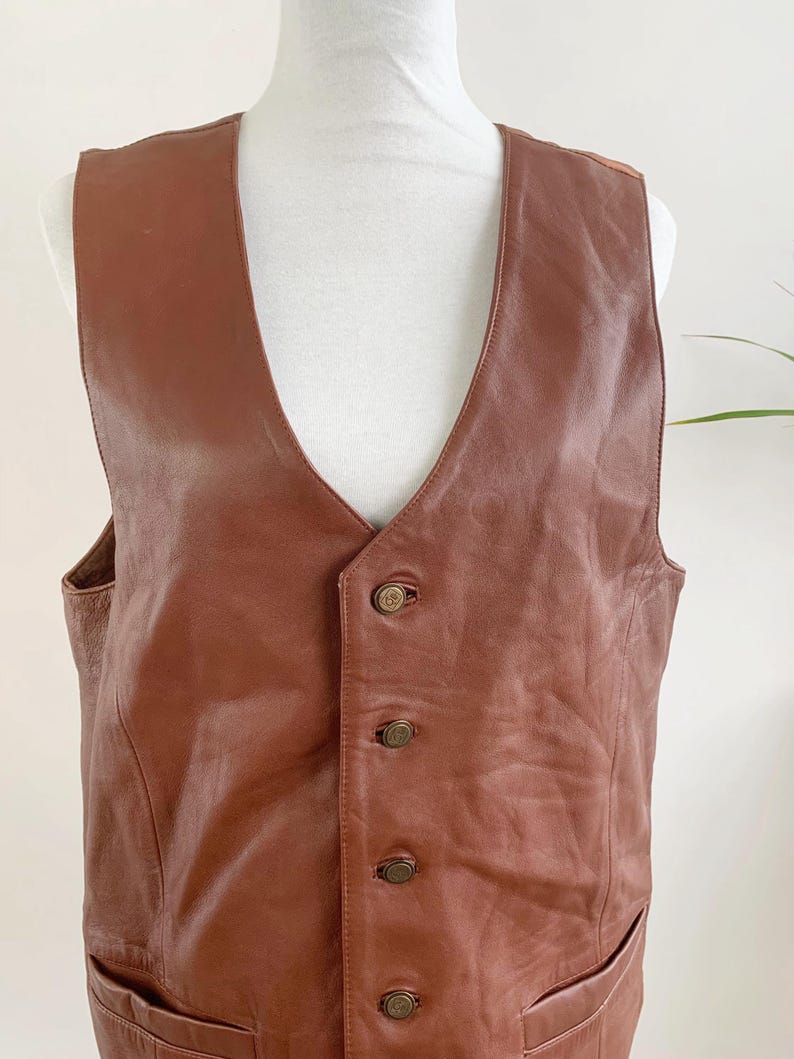 May include: A brown leather vest with a V-neck and four decorative buttons. The vest has two front pockets and is displayed on a white mannequin. The leather has a slightly distressed appearance.