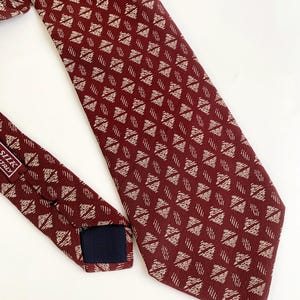 Könnte beinhalten: Eine bordeauxfarbene Seidenkrawatte mit einem sich wiederholenden Rautenmuster in einem helleren Beige. Die Krawatte liegt auf einer weißen Oberfläche und zeigt ihr Design und ihre Textur. Das Etikett lautet "SILK ITALY".