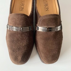 May include: A pair of brown suede loafers with a silver buckle detail. The loafers are made in Italy by the brand Peron.