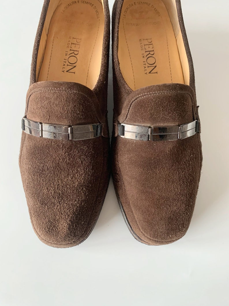 May include: A pair of brown suede loafers with a silver buckle detail. The loafers are made in Italy by the brand Peron.