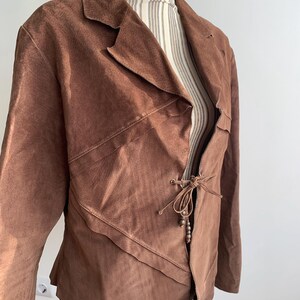 May include: A brown suede jacket with a tie closure and wooden beads. The jacket has a unique asymmetrical design and a relaxed fit.
