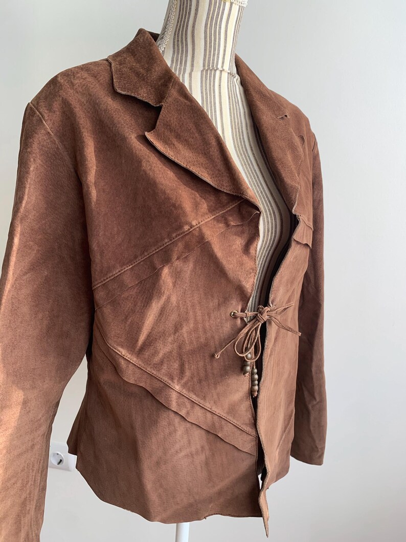 May include: A brown suede jacket with a tie closure and wooden beads. The jacket has a unique asymmetrical design and a relaxed fit.