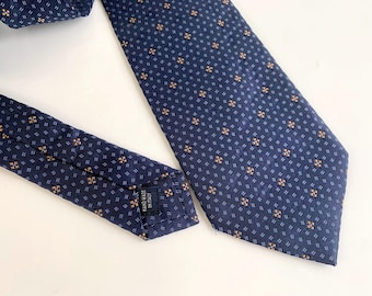 Könnte beinhalten: Eine marineblaue Krawatte mit einem sich wiederholenden Muster aus kleinen, braunen und weißen Blumenmustern. Die Krawatte ist aus glattem Stoff gefertigt und trägt ein Etikett "HAND MADE IN ITALY".