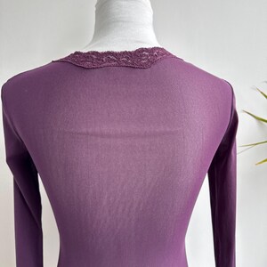 May include: A long-sleeved, plum-colored top with a delicate lace trim around the neckline. The fabric appears to be lightweight and form-fitting, with a subtle sheen. The top is displayed on a mannequin.