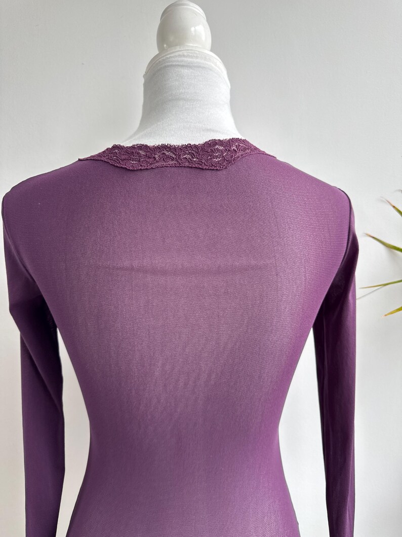 May include: A long-sleeved, plum-colored top with a delicate lace trim around the neckline. The fabric appears to be lightweight and form-fitting, with a subtle sheen. The top is displayed on a mannequin.