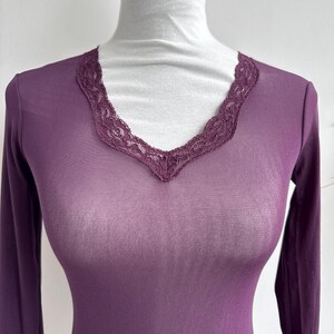 May include: Plum-colored long-sleeve top with a V-neckline trimmed with delicate lace. The top is made of a sheer, lightweight fabric. The garment is displayed on a white mannequin.