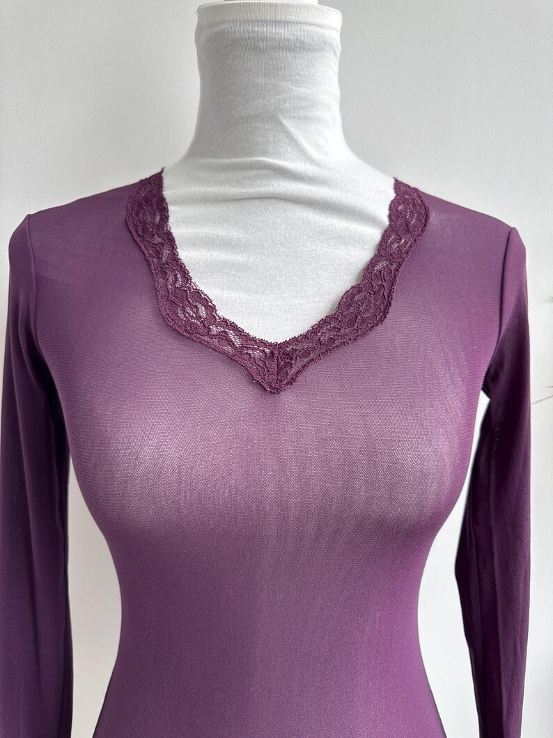 May include: Plum-colored long-sleeve top with a V-neckline trimmed with delicate lace. The top is made of a sheer, lightweight fabric. The garment is displayed on a white mannequin.