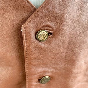 May include: Close-up of a brown leather vest with two brass-colored buttons. The buttons have a decorative emblem. The vest has a V-neck and is stitched with a matching brown thread. The leather has a smooth texture.