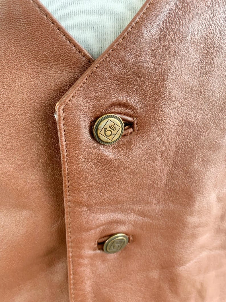 May include: Close-up of a brown leather vest with two brass-colored buttons. The buttons have a decorative emblem. The vest has a V-neck and is stitched with a matching brown thread. The leather has a smooth texture.