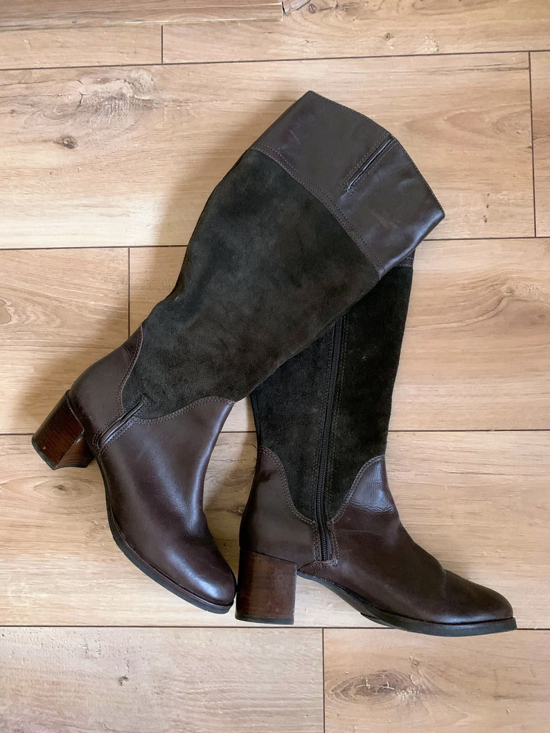 May include: A pair of brown leather knee-high boots with a suede panel on the front and back. The boots have a block heel and a zipper on the inside.