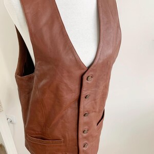 May include: A brown leather vest with a V-neck and a button-up front. The vest has two front pockets and is displayed on a white mannequin. The leather appears to be a medium brown color.