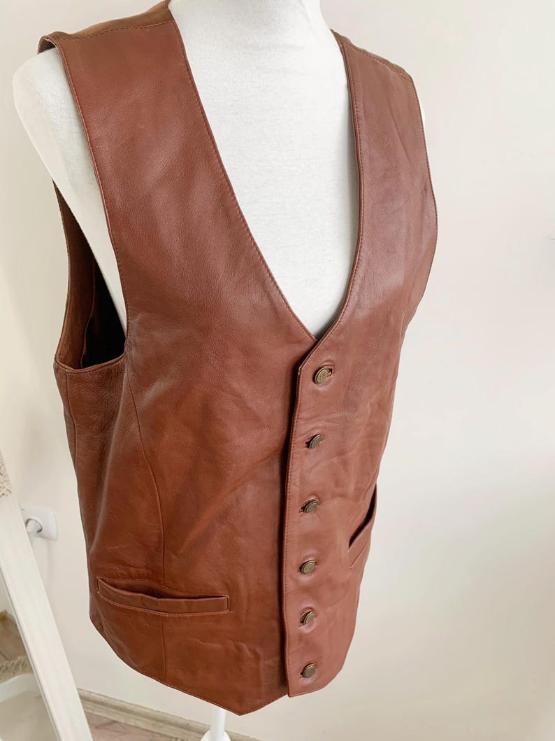 May include: A brown leather vest with a V-neck and a button-up front. The vest has two front pockets and is displayed on a white mannequin. The leather appears to be a medium brown color.