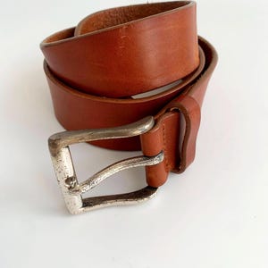 May include: A rolled, brown leather belt with a silver-colored buckle. The belt is a medium brown color and appears to be made of high-quality leather. The buckle is rectangular and has a worn appearance.