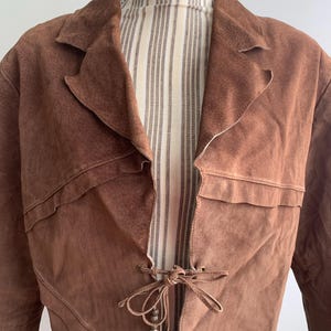 May include: A brown suede jacket with a tie closure and beaded accents. The jacket has a long, flowing silhouette and a relaxed fit.