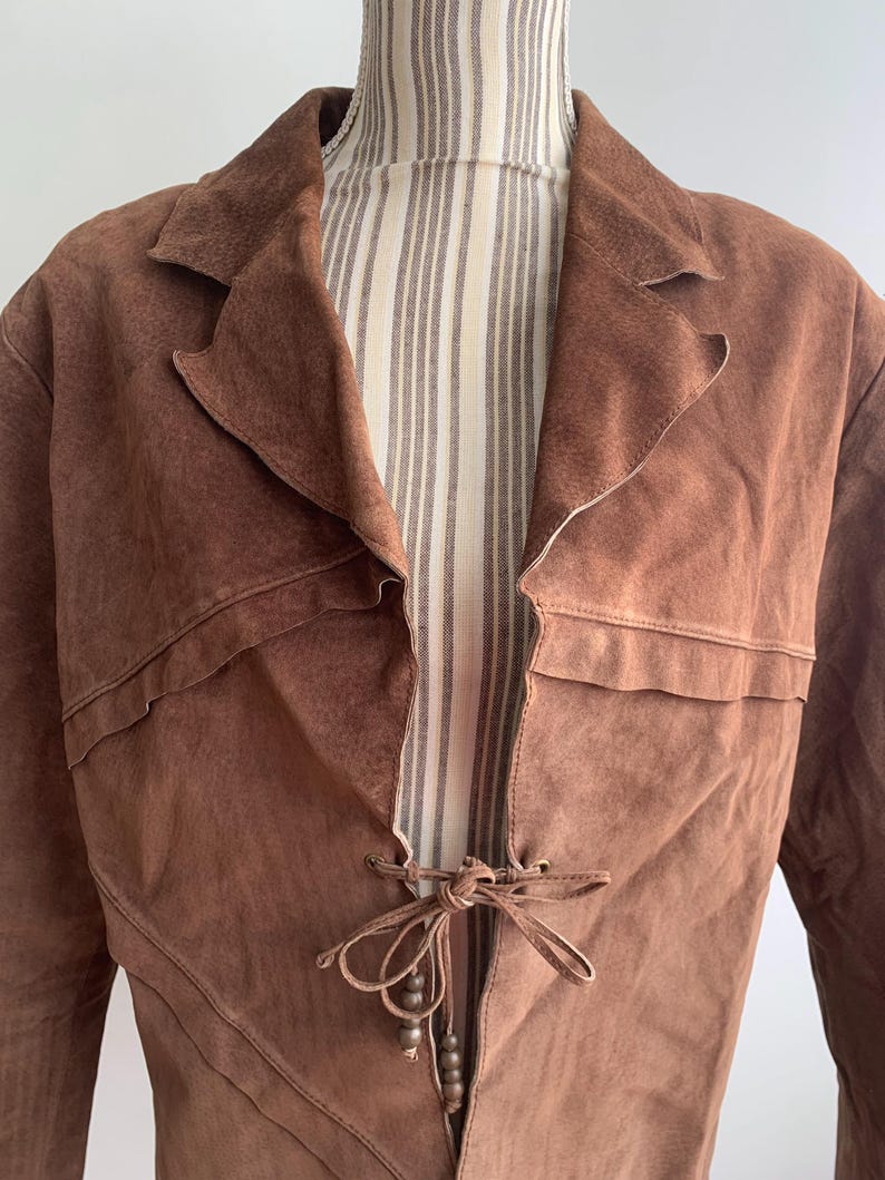May include: A brown suede jacket with a tie closure and beaded accents. The jacket has a long, flowing silhouette and a relaxed fit.