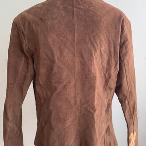 May include: A brown suede jacket with a collared neckline and long sleeves. The jacket has a fitted silhouette and a slightly flared hem.