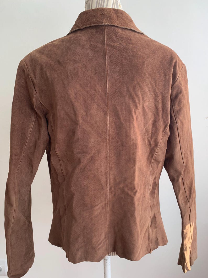 May include: A brown suede jacket with a collared neckline and long sleeves. The jacket has a fitted silhouette and a slightly flared hem.