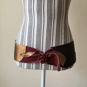 May include: A wide leather belt with a unique design featuring burgundy, gold, and brown leather panels. The belt has a buckle with a decorative design.