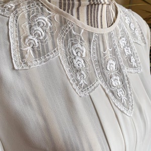 Puede incluir: Una blusa blanca con un delicado cuello de encaje con un dise&ntilde;o floral. El cuello es festoneado y tiene una calidad transparente, casi transl&uacute;cida.