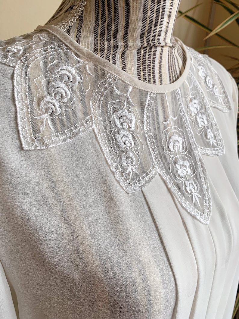 Puede incluir: Una blusa blanca con un delicado cuello de encaje con un dise&ntilde;o floral. El cuello es festoneado y tiene una calidad transparente, casi transl&uacute;cida.