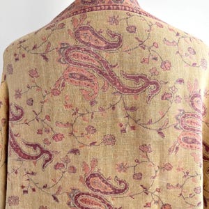 May include: A beige shawl with a paisley pattern in shades of pink and burgundy. The shawl is draped over a form, showcasing the intricate floral and paisley design. The fabric appears soft and lightweight.