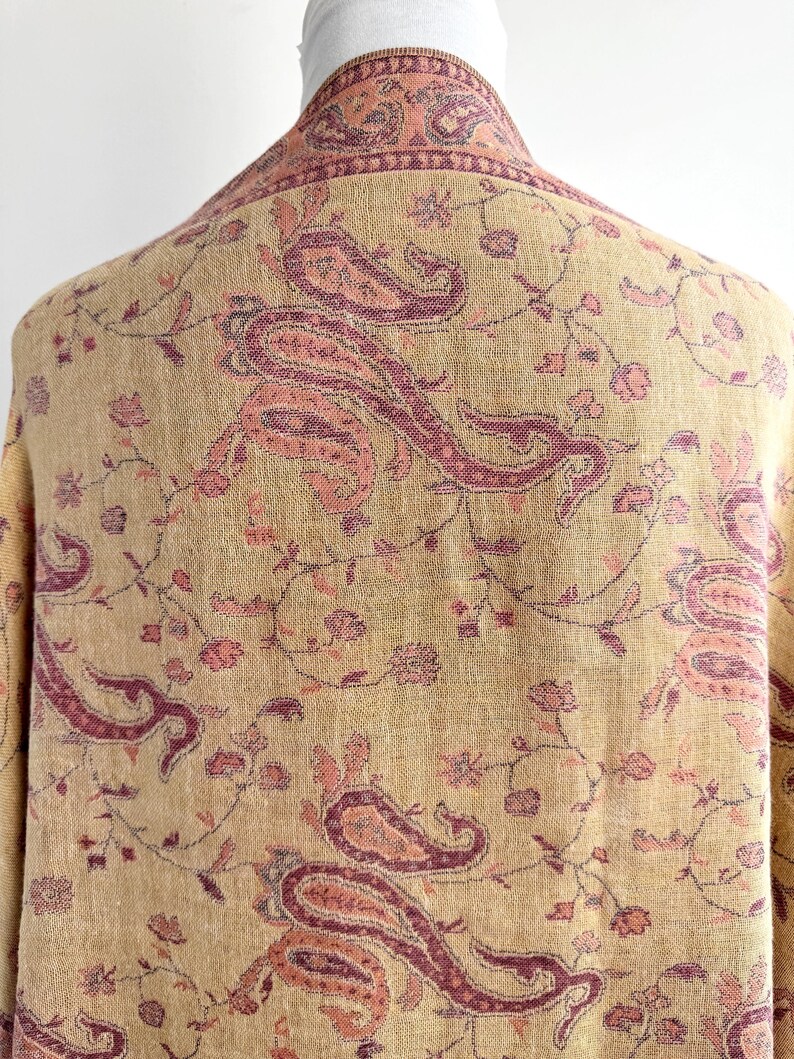 May include: A beige shawl with a paisley pattern in shades of pink and burgundy. The shawl is draped over a form, showcasing the intricate floral and paisley design. The fabric appears soft and lightweight.