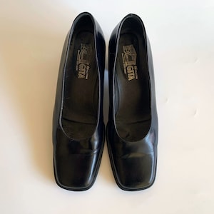 May include: A pair of black leather pumps with a square toe. The shoes have a low, block heel and the brand name "GITA" is visible inside the shoe. The shoes are displayed on a white surface.