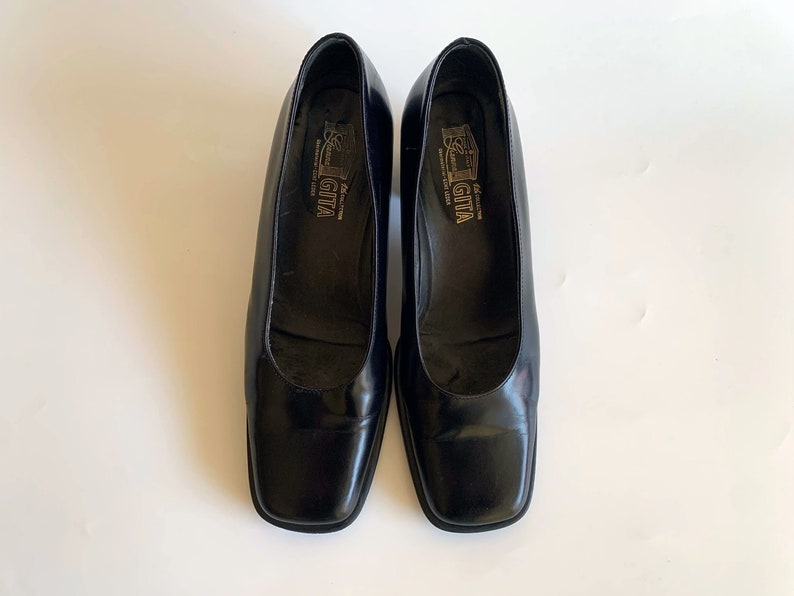 May include: A pair of black leather pumps with a square toe. The shoes have a low, block heel and the brand name "GITA" is visible inside the shoe. The shoes are displayed on a white surface.