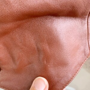 May include: Close-up of a brown leather item, showing the texture and color variations. The leather has a smooth, slightly glossy finish with subtle imperfections. The edges of the item are visible, suggesting a garment or accessory.