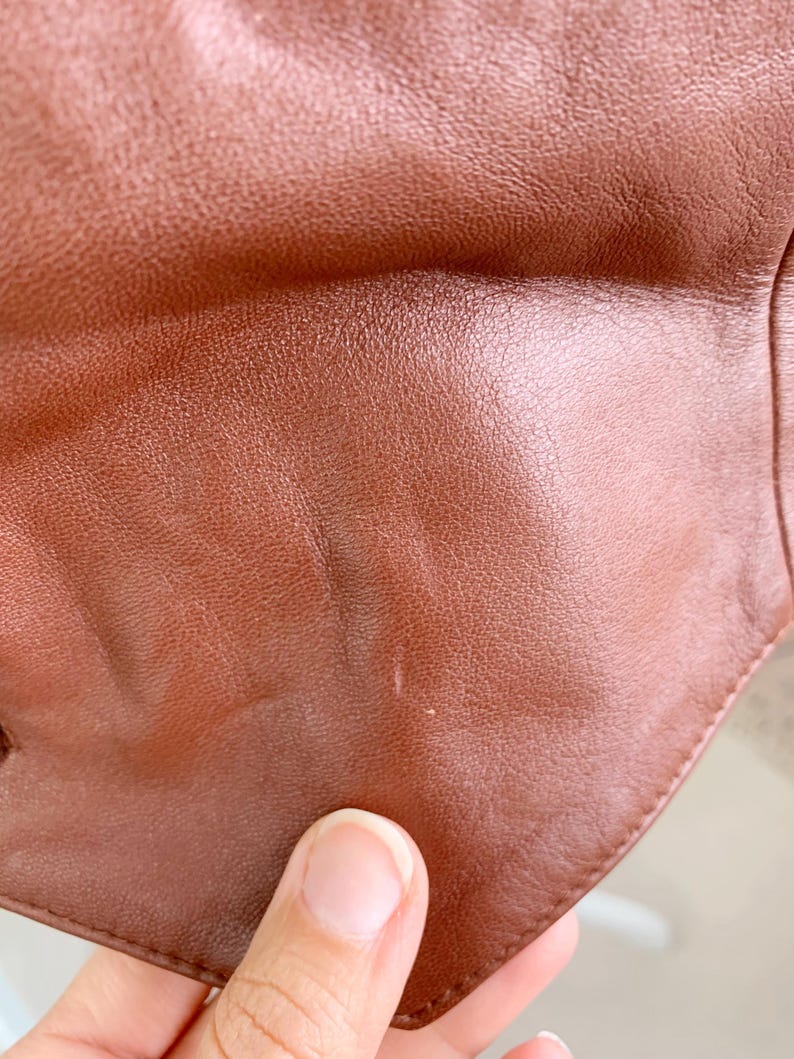 May include: Close-up of a brown leather item, showing the texture and color variations. The leather has a smooth, slightly glossy finish with subtle imperfections. The edges of the item are visible, suggesting a garment or accessory.