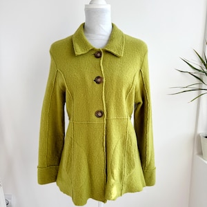 Könnte beinhalten: Eine hellgrüne Jacke mit Kragen und drei braunen Knöpfen. Die langärmlige Jacke hat eine taillierte Passform und wird auf einer weißen Schaufensterpuppe präsentiert. Die Nähte der Jacke sind sichtbar und tragen zu ihrem Design bei.