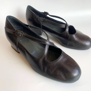 Pu&ograve; includere: Un paio di scarpe con tacco in pelle nera con un design a cinturino incrociato e una chiusura a fibbia.
