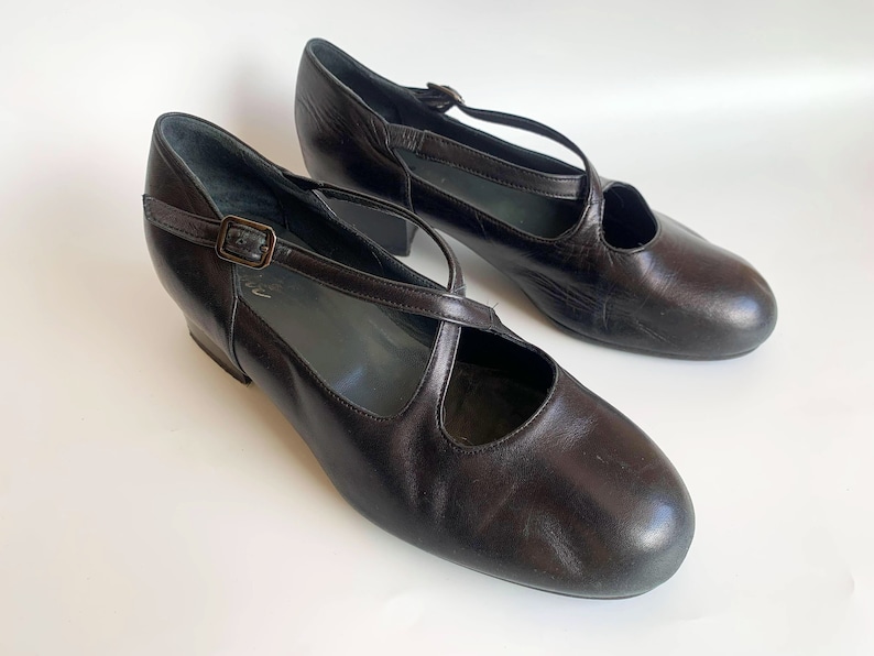 Pu&ograve; includere: Un paio di scarpe con tacco in pelle nera con un design a cinturino incrociato e una chiusura a fibbia.