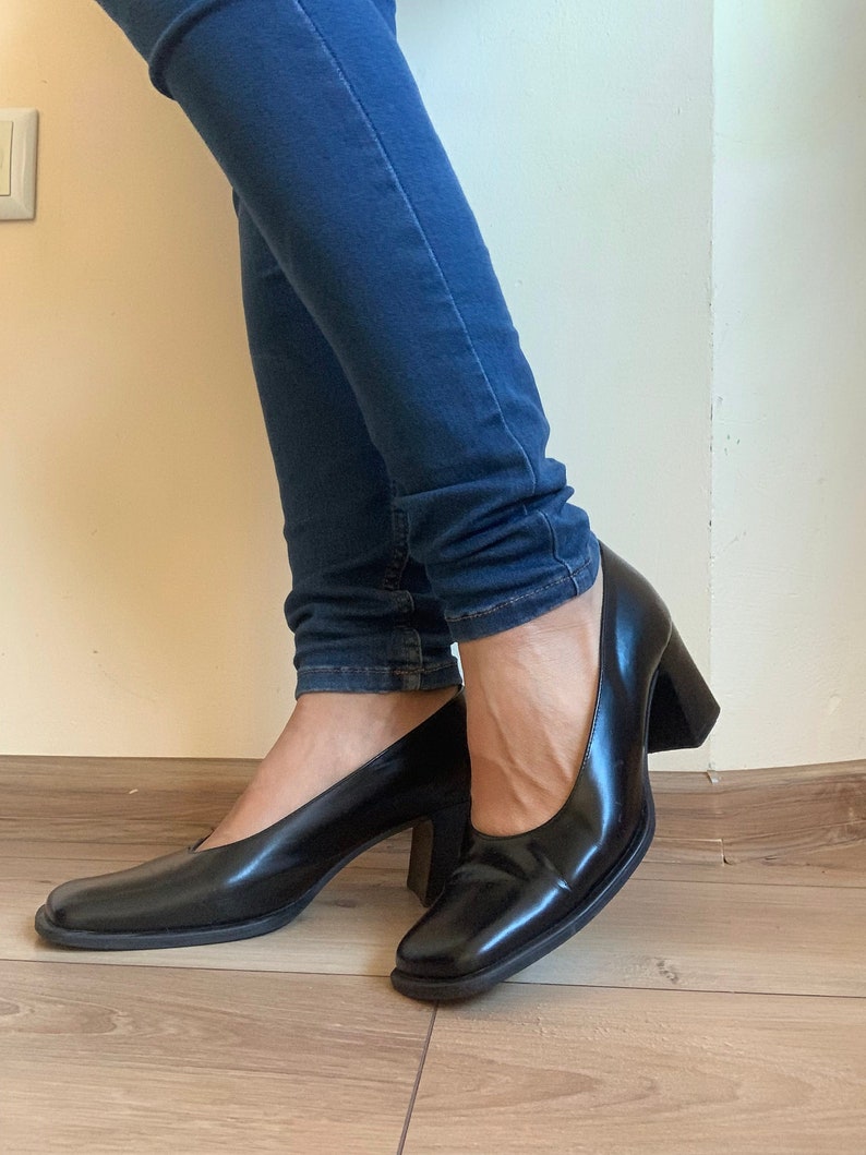 May include: Black leather pumps with a square toe and a block heel. The shoes have a glossy finish and are worn with blue jeans. The heels appear to be approximately 3 inches high.