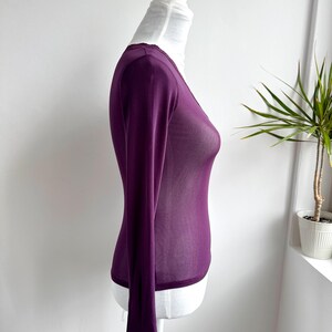 May include: A long-sleeved, deep purple top with a V-neck and delicate lace trim at the cuffs and neckline. The sheer fabric drapes over a white mannequin, showcasing the garment's form and design. The top is a stylish addition to any wardrobe.